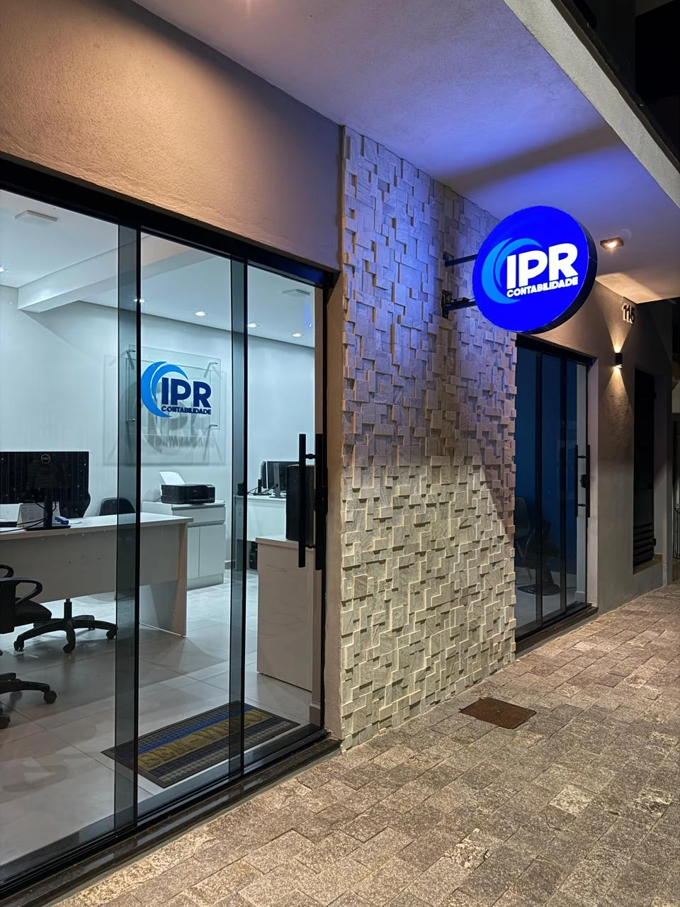 Fachada real da IPR Contabilidade, com portas de vidro, revestimento de pedra e letreiro circular azul iluminado